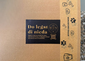 Versandkarton von Nature's Gold mit bayerischem Aufkleber „Do legst di nieda“ und Paketband mit Hundemotiven.