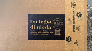 Versandkarton von Nature's Gold mit bayerischem Aufkleber „Do legst di nieda“ und Paketband mit Hundemotiven.